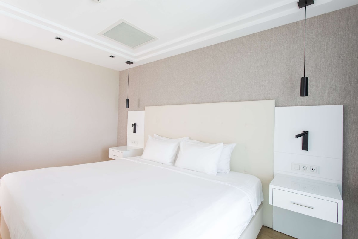 Ein modernes Hotelzimmer mit einem großen Bett, einem Nachttisch, einem an der Wand montierten Fernseher und einem kleinen Schreibtisch mit Stuhl.