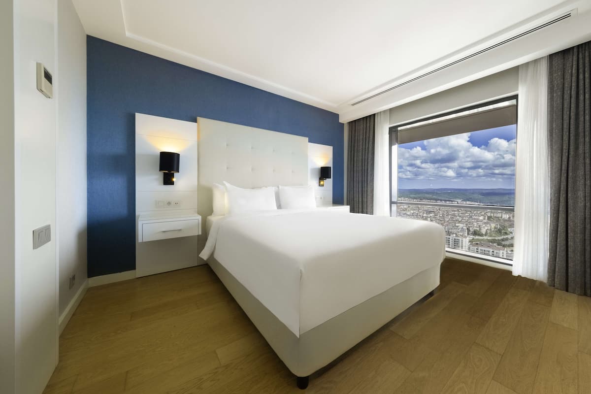 Ein Hotelzimmer mit einem großen Bett, einem Nachttisch, einer Wandlampe und einem Fenster mit Blick auf die Stadt.