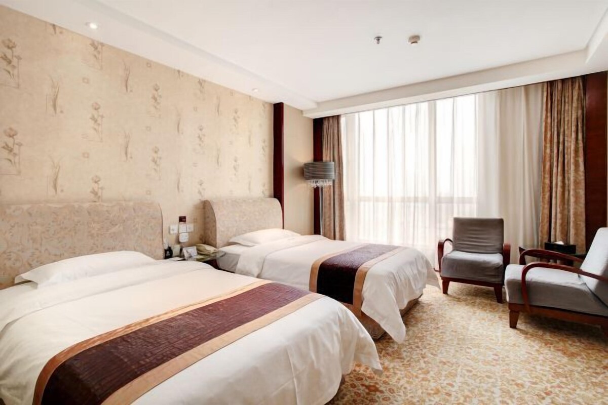 Hotelkamer met twee bedden, een zithoek met een stoel, een lamp en een groot raam met gordijnen.