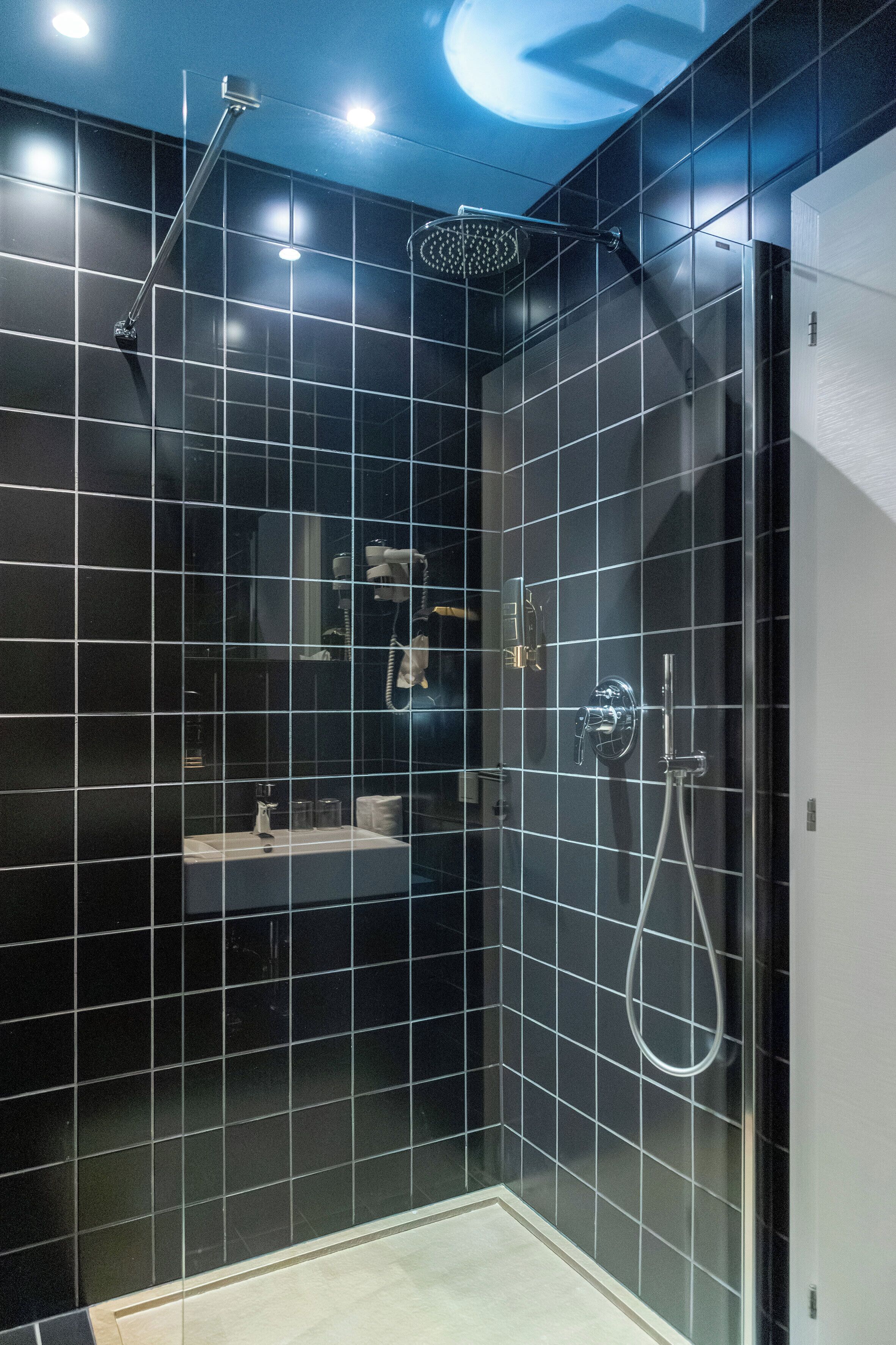 Une salle de bain moderne avec des carreaux noirs, une douche à l’italienne et un petit meuble-lavabo avec un miroir.