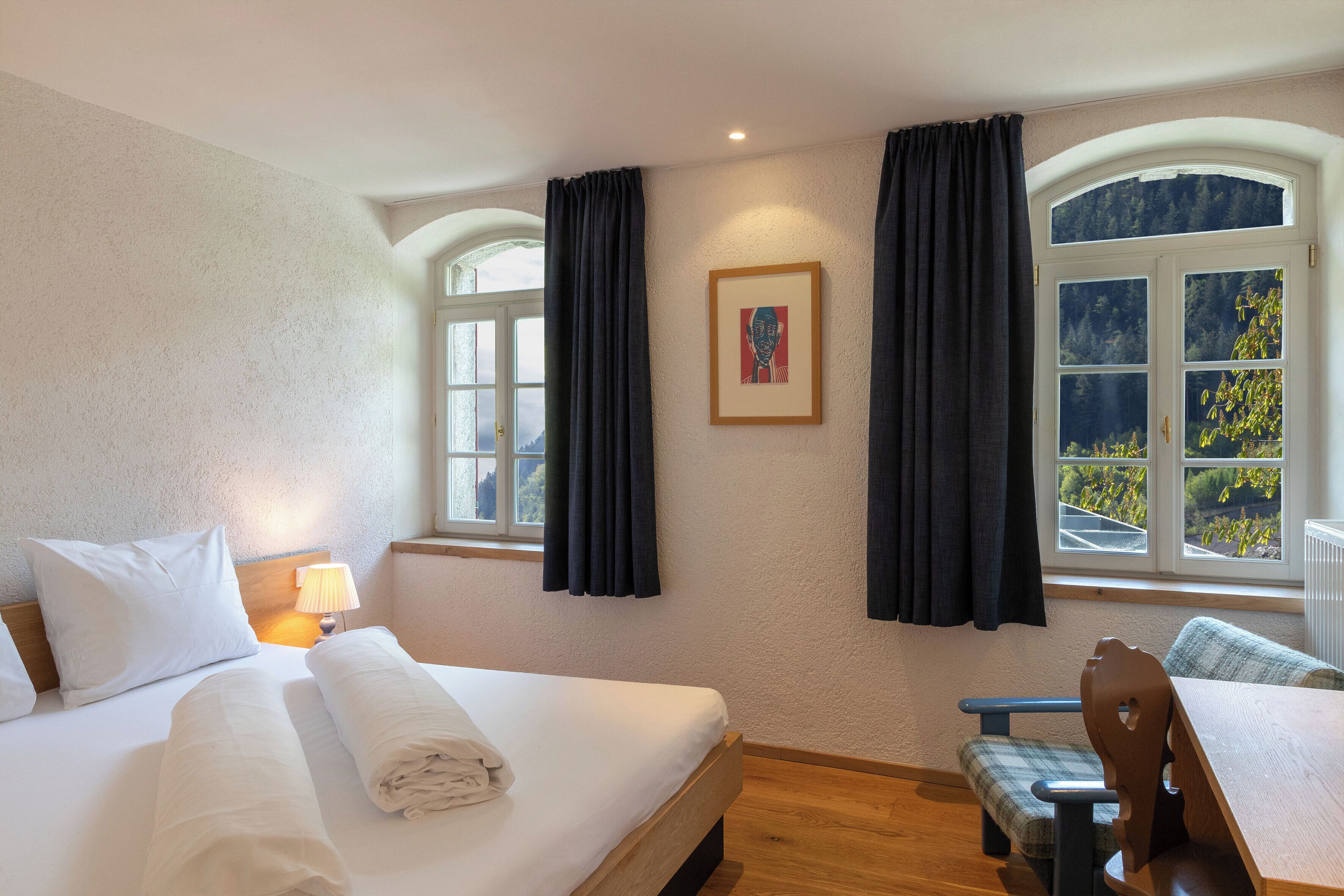 Una habitación de hotel con cama, una banca, una mesa, una ventana con cortinas y un cuadro en la pared.