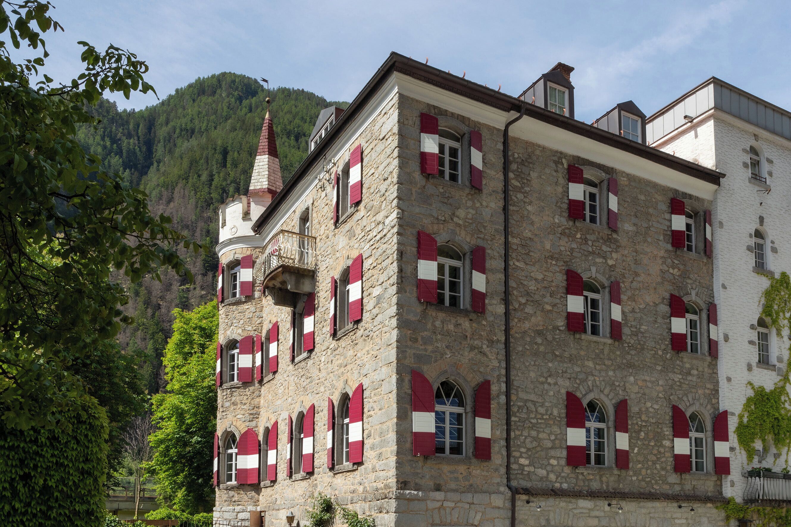 A stone building with red and white shutters, a turret, and a mountainous backdrop.
