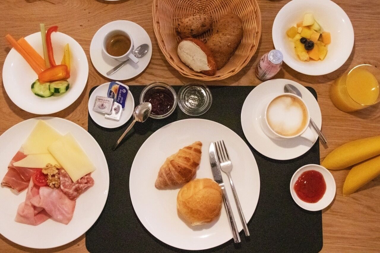 Una mesa de desayuno con platos de comida, incluyendo croissants, queso y frutas, junto con tazas de café y jugo.
