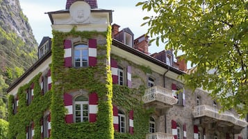 Um edifício histórico com paredes cobertas de hera, uma torre imponente e venezianas vermelhas e brancas.