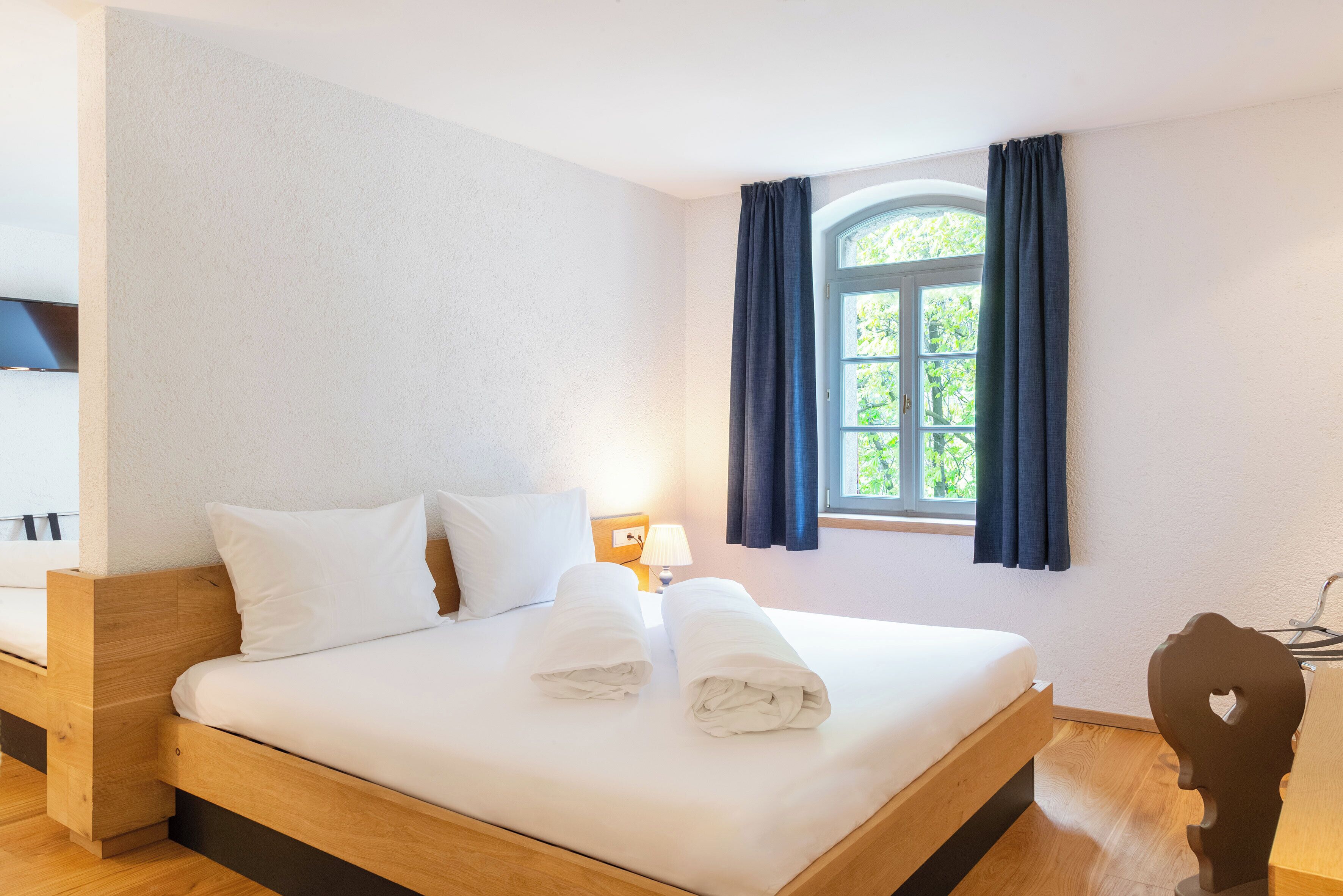 Een net opgemaakt bed met witte lakens en twee handdoeken, een nachtkastje met een lamp, een raam met gordijnen en een houten hoofdbord.
