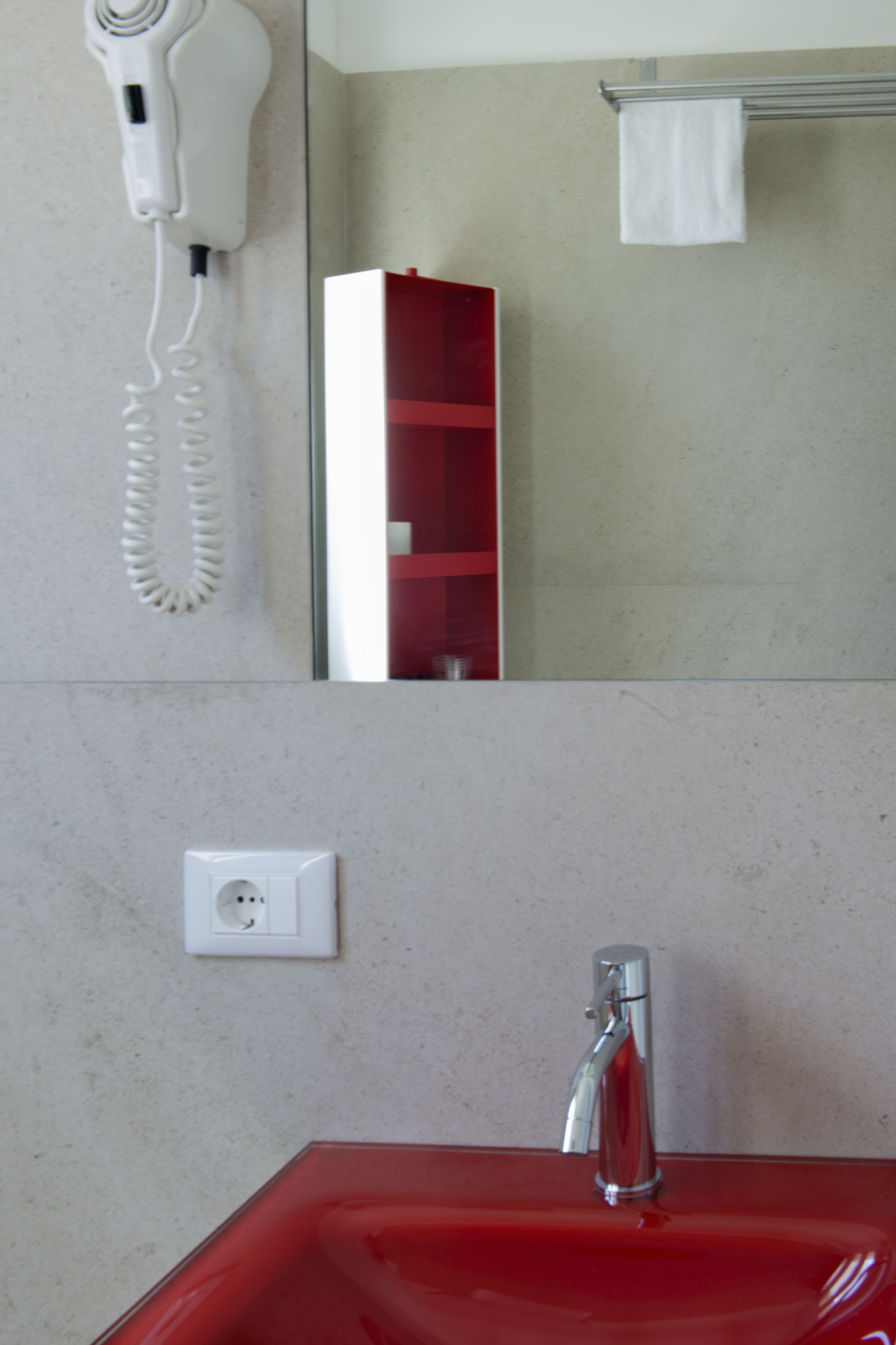 Un baño con un lavabo rojo, un secador de pelo blanco montado en la pared, y un espejo que refleja una repisa roja y una toalla blanca.