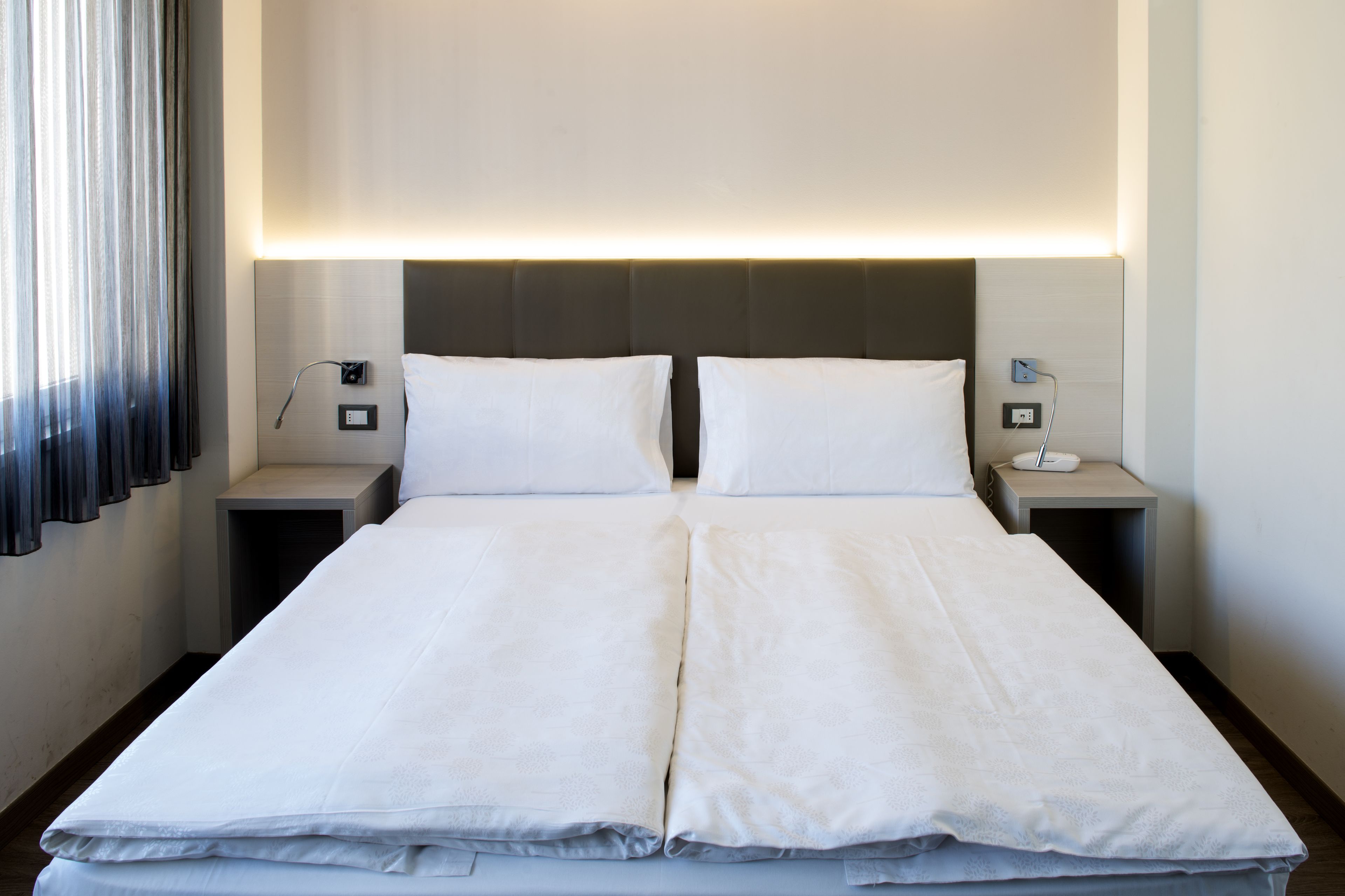 Ein Doppelbett mit weißer Bettwäsche, zwei Nachttische mit Lampen und ein Kopfteil mit integrierter Beleuchtung.