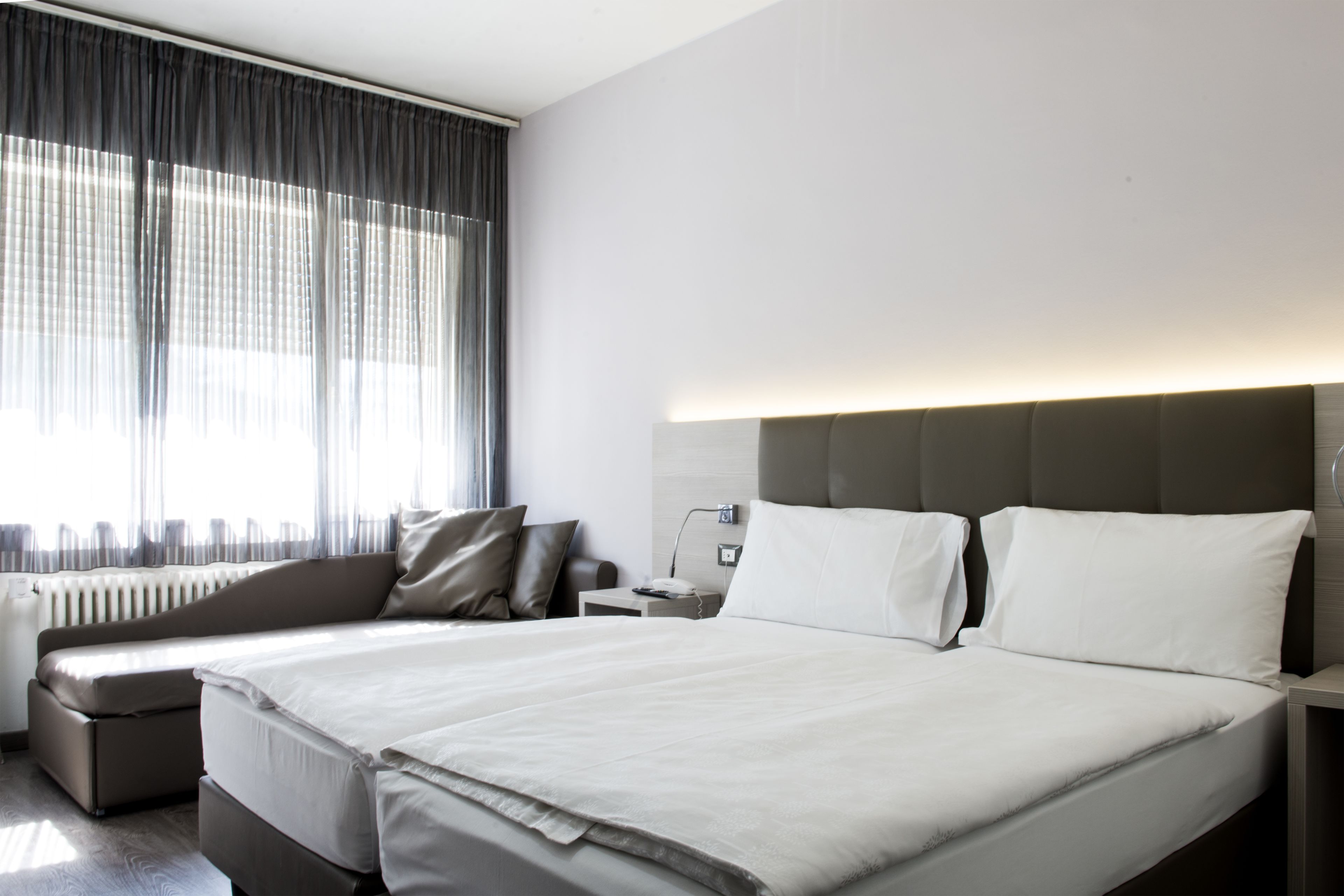 Una habitación de hotel moderna con una cama grande, un sofá, una mesita de noche y una ventana con cortinas traslúcidas.