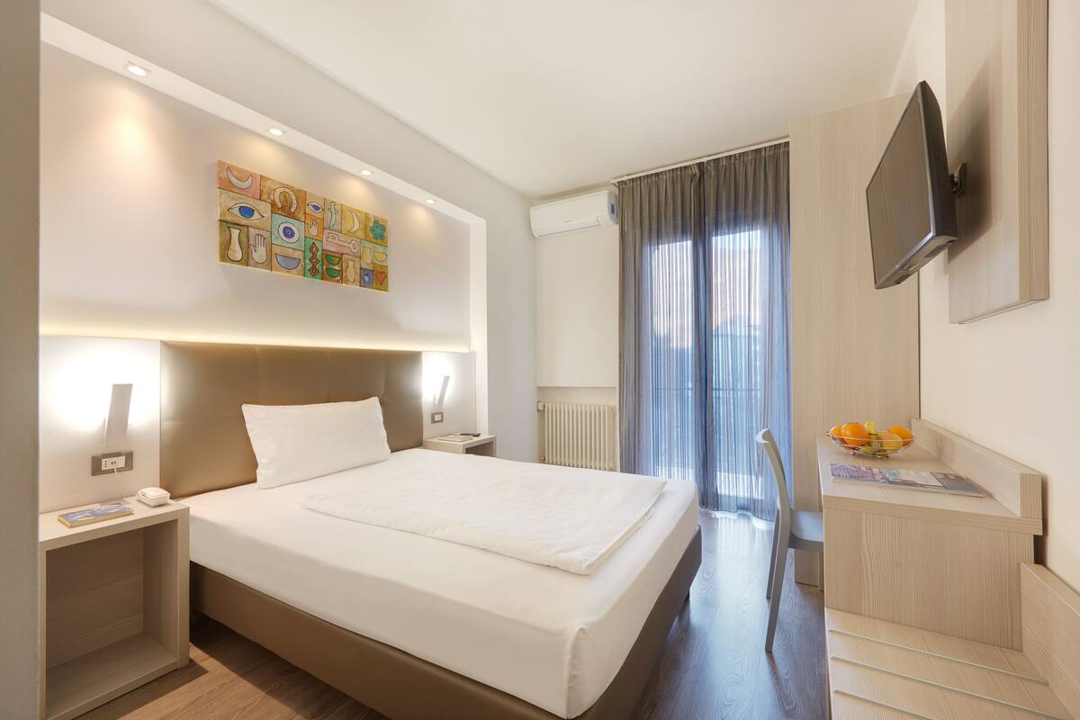 Habitación de hotel moderna con una cama grande, mesitas de noche, un televisor de pantalla plana, un cuadro en la pared y vistas al exterior a través de una ventana.