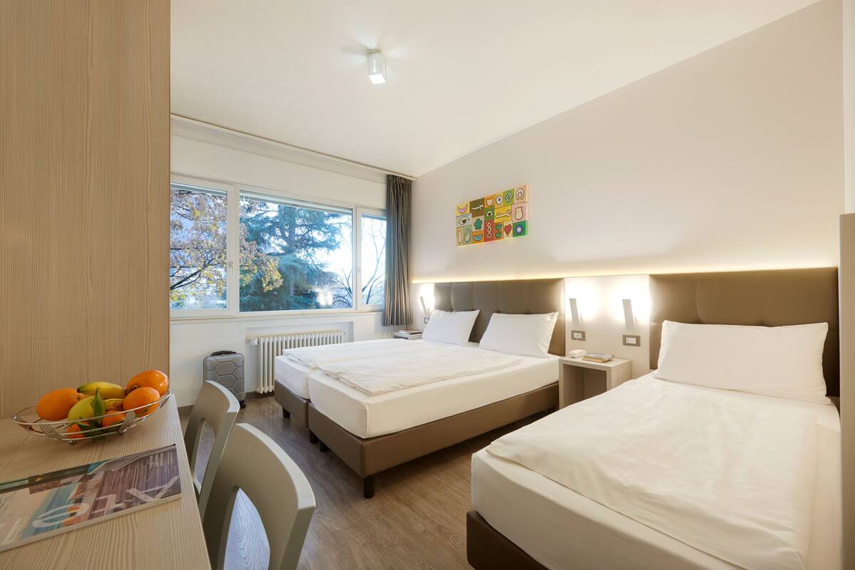 Habitación de hotel con dos camas, un escritorio con frutero, un radiador y una ventana con vista a árboles.