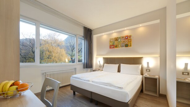 Una habitación de hotel moderna con una cama grande, mesitas de noche, un escritorio con frutero y una ventana con vistas a árboles.