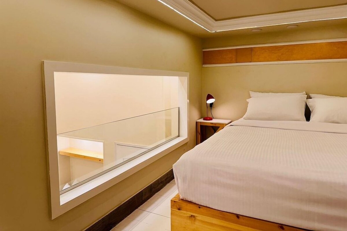 A bedroom with a large bed, a bedside table with a lamp, and a window with a view of the outside.