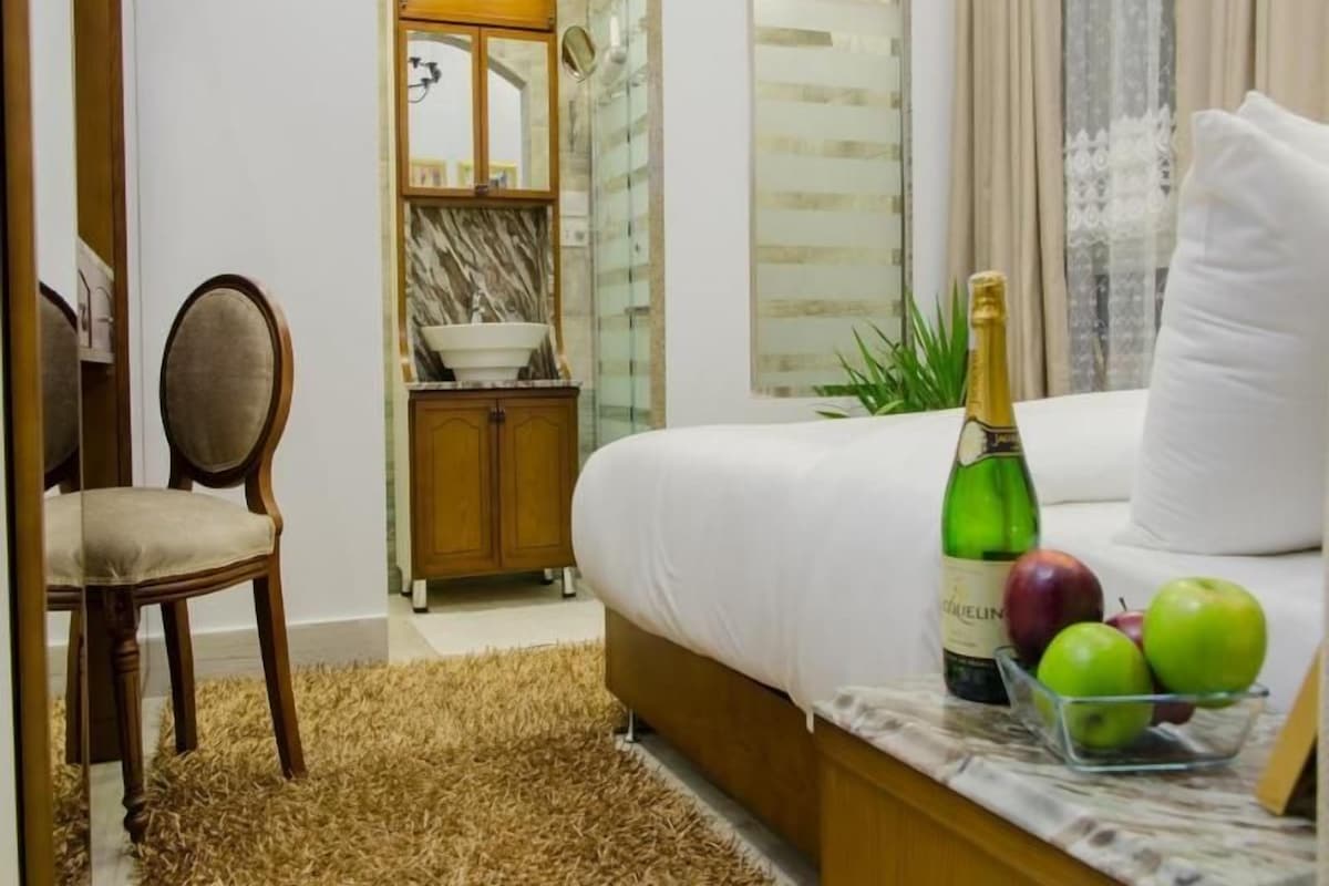A hotel room with a bed, a chair, a small table with a bottle and apples, and a bathroom visible in the background.