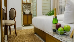 A hotel room with a bed, a wooden chair, a small table with a bottle of champagne and apples, a mirror, and a bathroom visible through an archway. - Paris Hotel (Cairo)