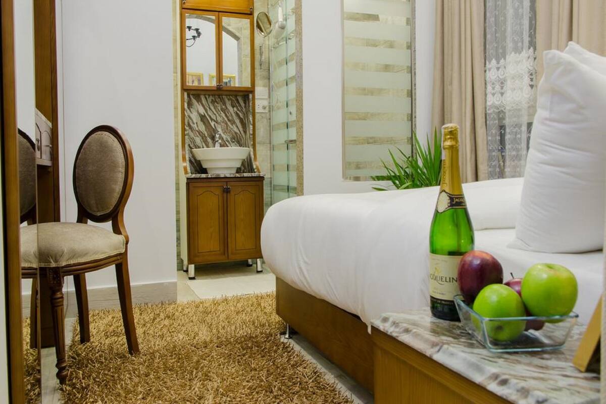 Ein Hotelzimmer mit einem Bett, einem Holzstuhl, einem kleinen Tisch mit Champagner und Äpfeln, einem Spiegel und einem durch einen Bogen sichtbaren Badezimmer.