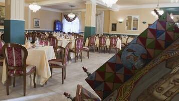A banquet hall with multiple tables and chairs, a chandelier, and a large, colorful mural on the wall.