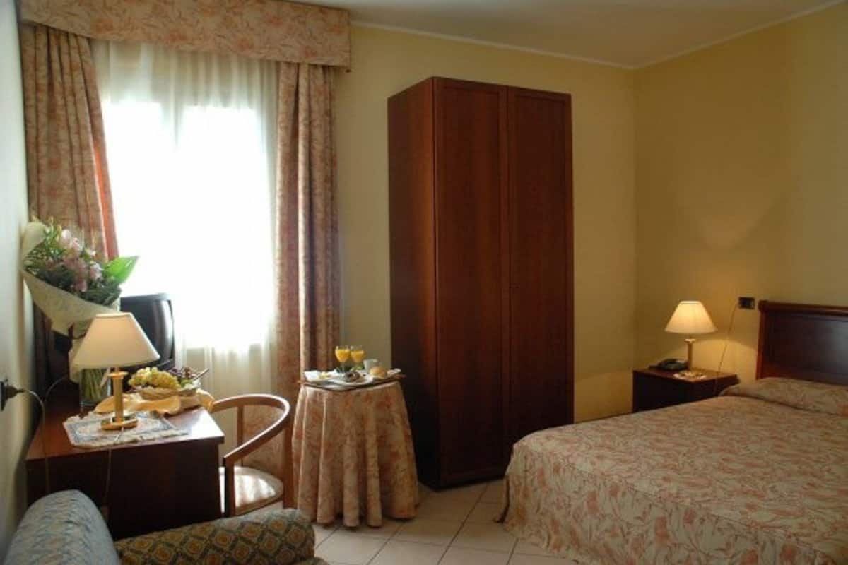 Una habitación de hotel con una cama, un escritorio con lámpara, una silla, un armario y una mesita con un jarrón de flores.