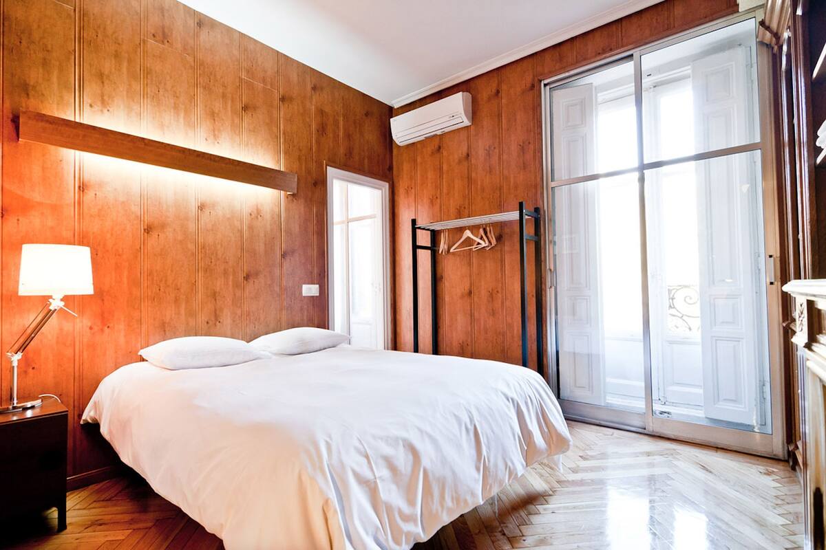 Un dormitorio con pared de madera, una cama, una mesita de noche, una lámpara, una ventana y un gran ventanal corredero.