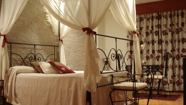 A four-poster canopy bed with a white bedspread and red pillows, a small table with a vase of flowers, and a chair. - El Castillo (Ponferrada)