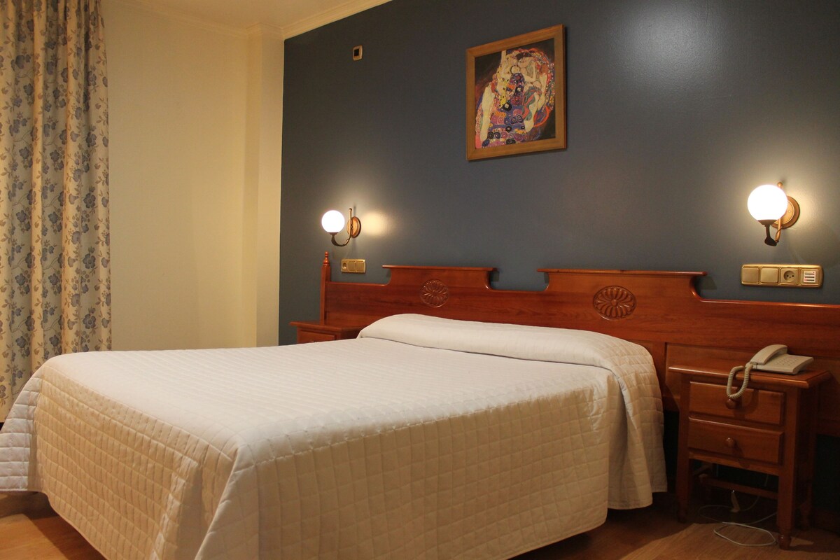 Una cama doble con cabecera de madera, dos lámparas de noche, una mesita con teléfono y un cuadro en la pared.