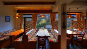 Un comedor con bancos y mesas de madera, ventanas amplias con cortinas, y lámparas colgantes.