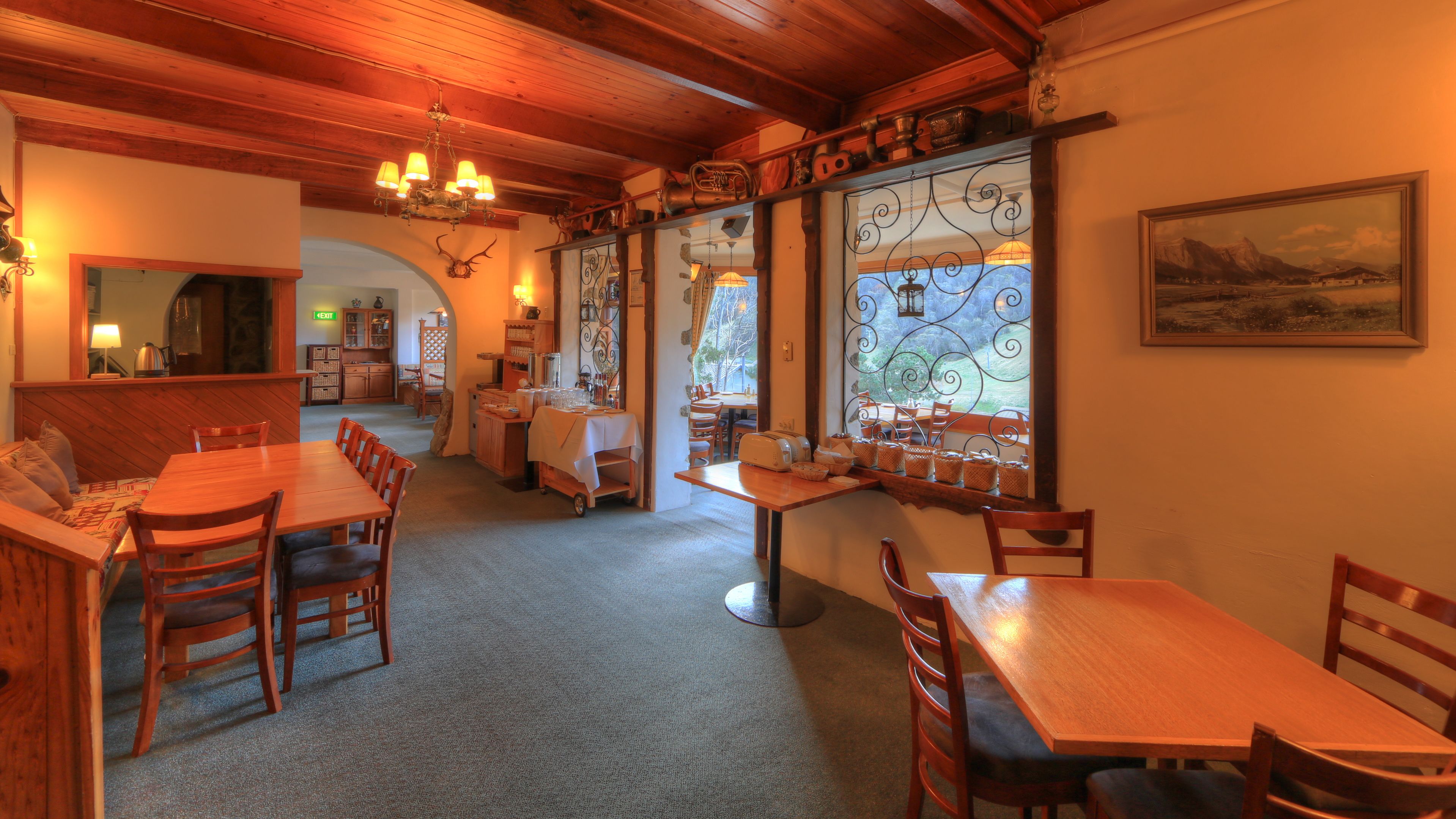 A cozy dining area with wooden tables and chairs, a chandelier, and a view of the outdoors through a window with decorative ironwork.