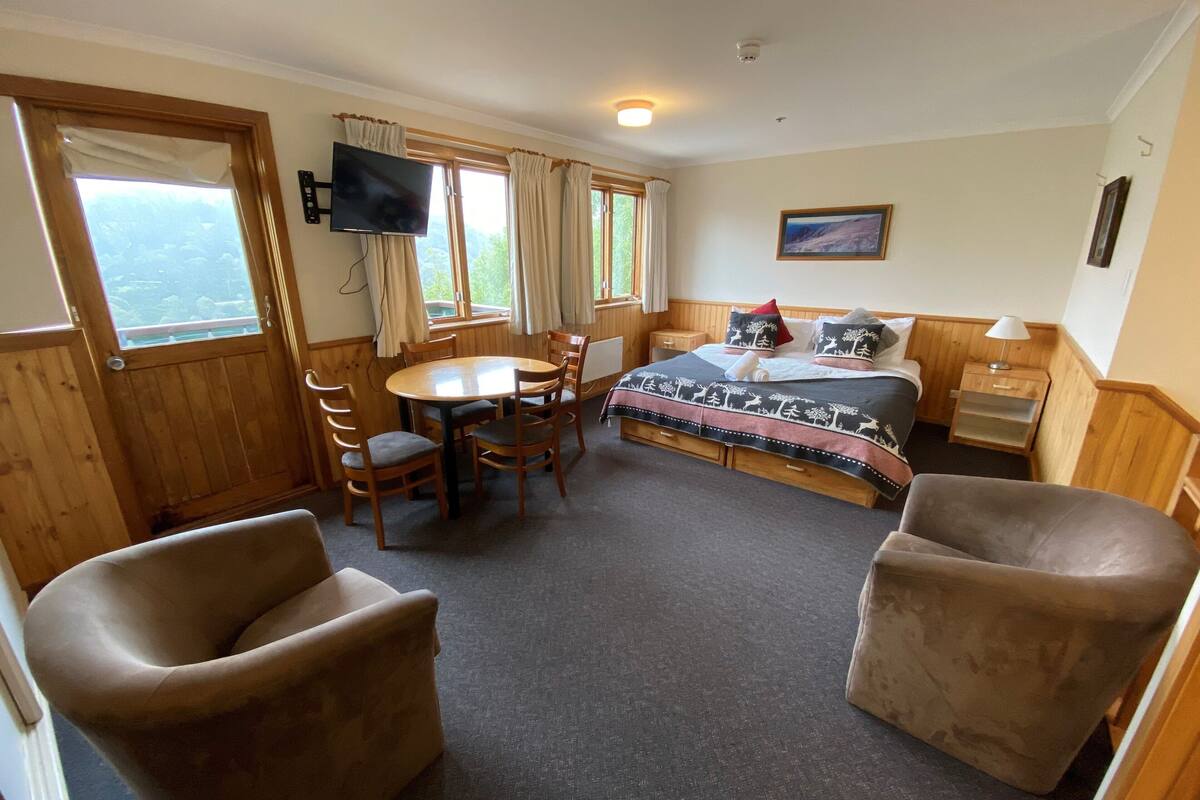 A room with a bed, a TV, a dining table with chairs, and a window with curtains.