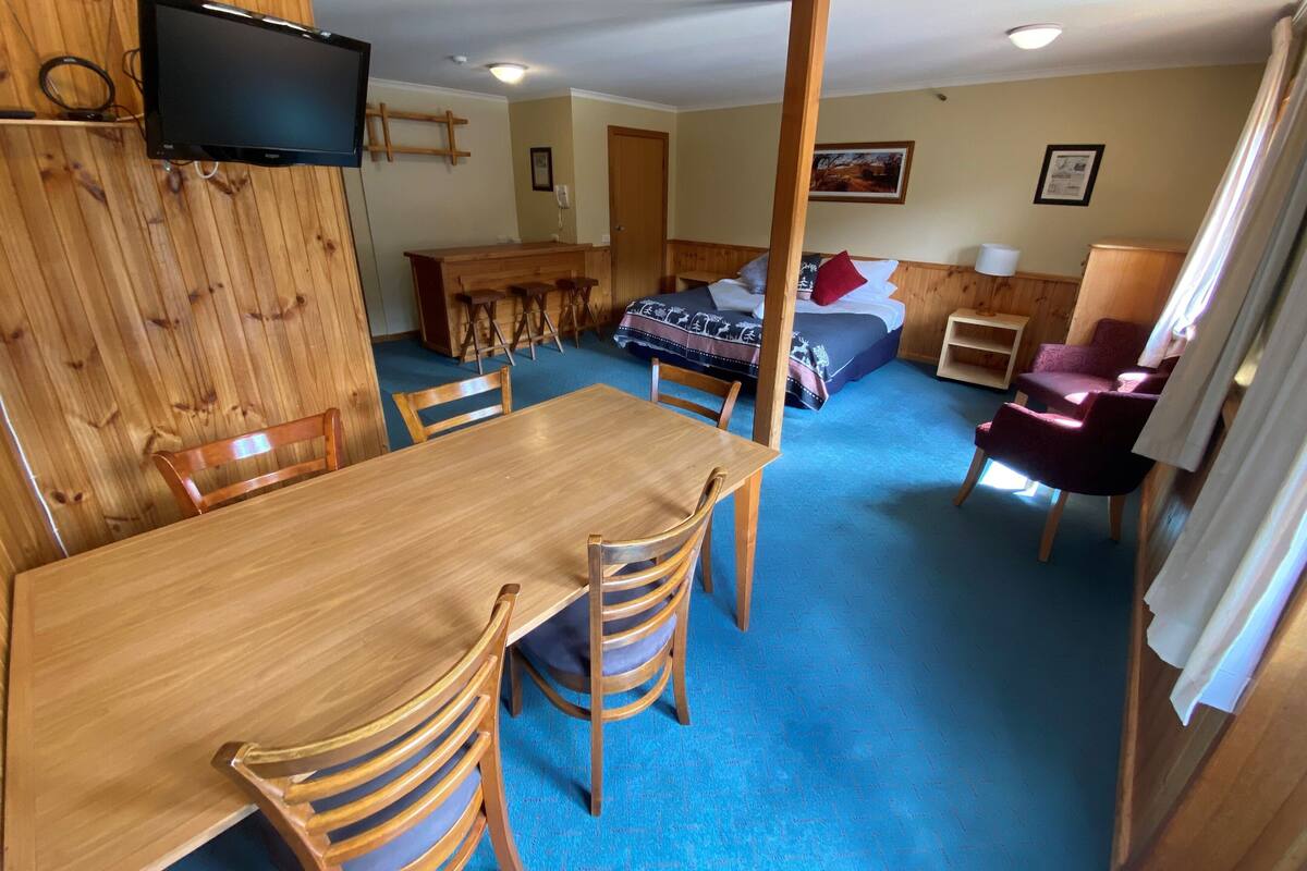 A room with a wooden dining table and chairs, a bed with a blue blanket, a TV mounted on the wall, and a wooden bar area with stools.