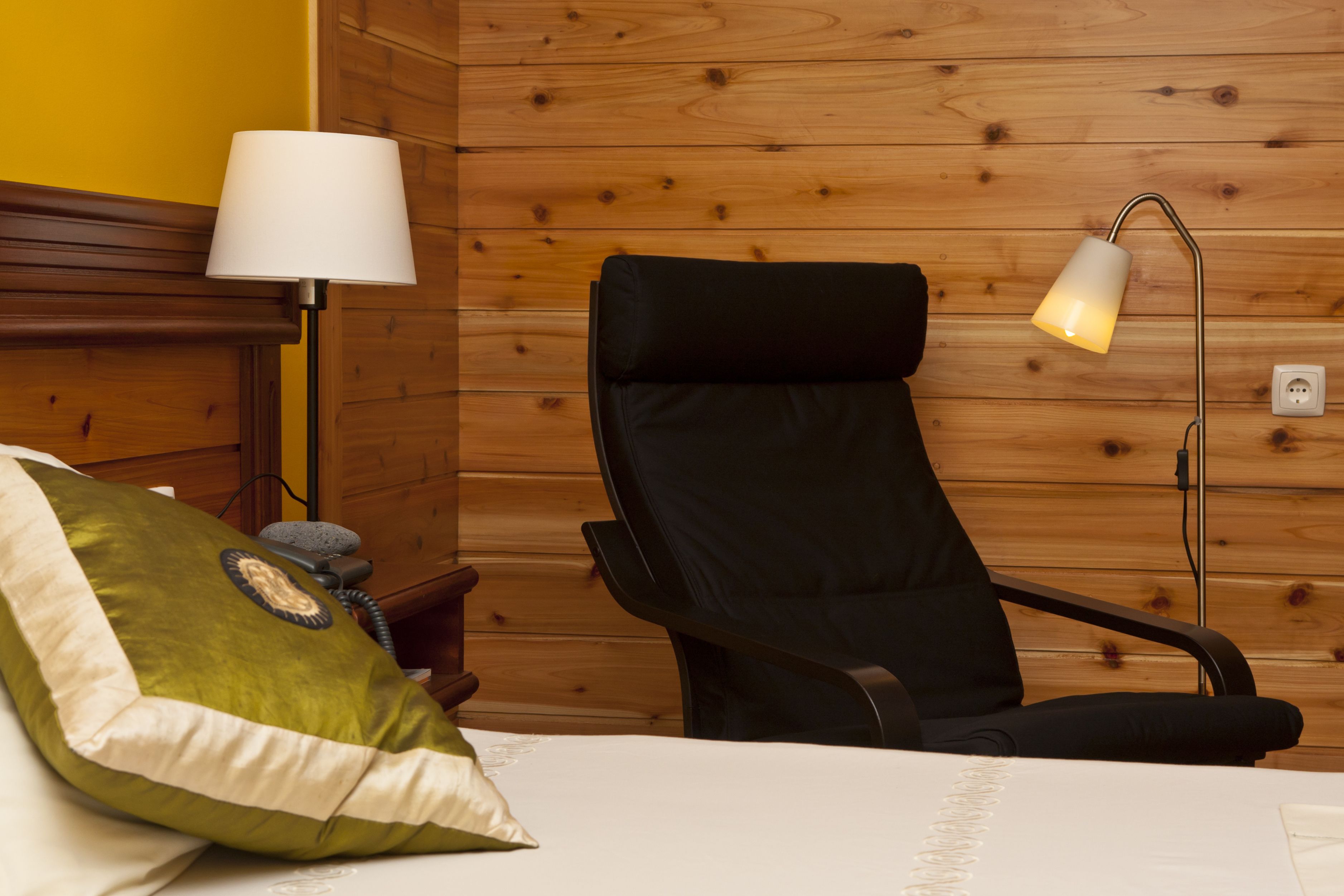Ein Hotelzimmer mit Holzwand, einem schwarzen Massagesessel, einem weißen Bett mit grünem Kopfkissen und einer Stehlampe.