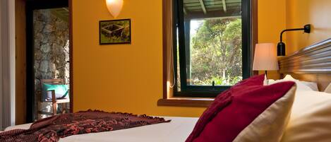 A bedroom with a bed, a window with a view of greenery, a wall-mounted lamp, and a stone wall.