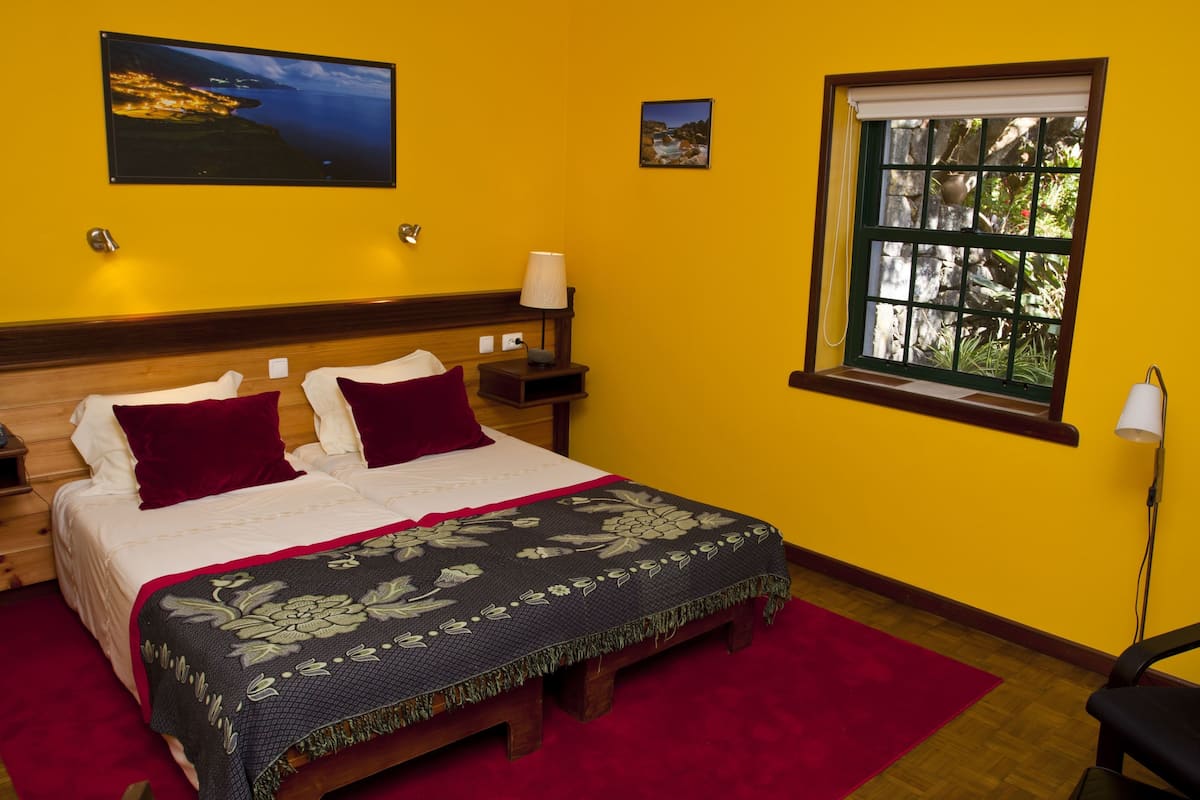 A hotel room with a wooden bed, a red rug, a nightstand with a lamp, and a window with a view of trees.