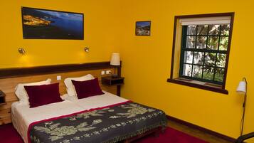 A hotel room with a wooden bed, a red rug, a nightstand with a lamp, and a window with a view of trees.