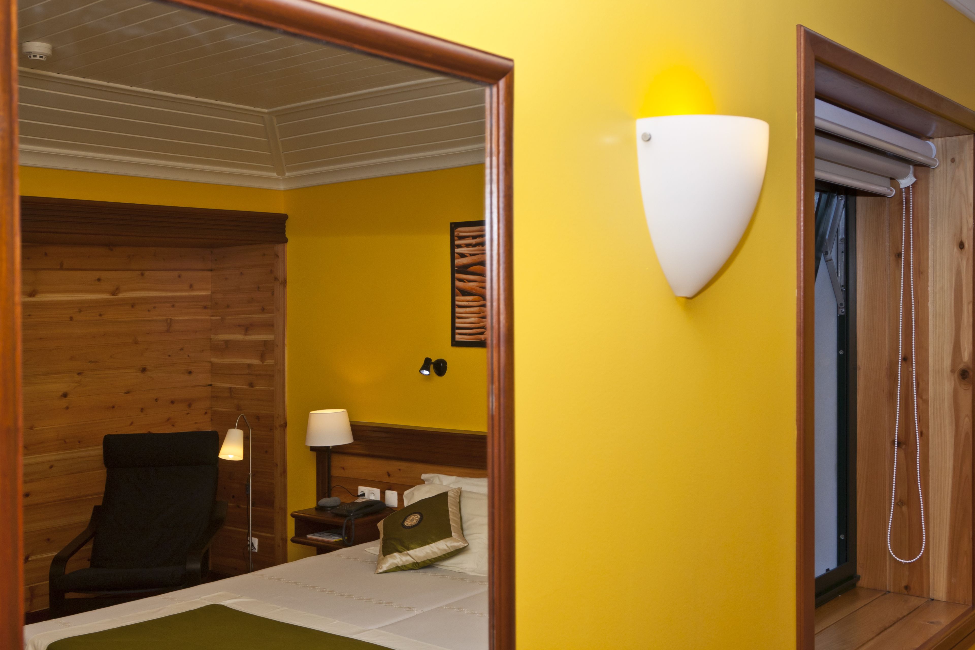 A hotel room with a wooden headboard, a bed with a green throw, a nightstand with a lamp, and a wall-mounted light fixture.