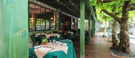 An outdoor dining area with tables covered in green tablecloths, wooden flooring, and a tree in the background.