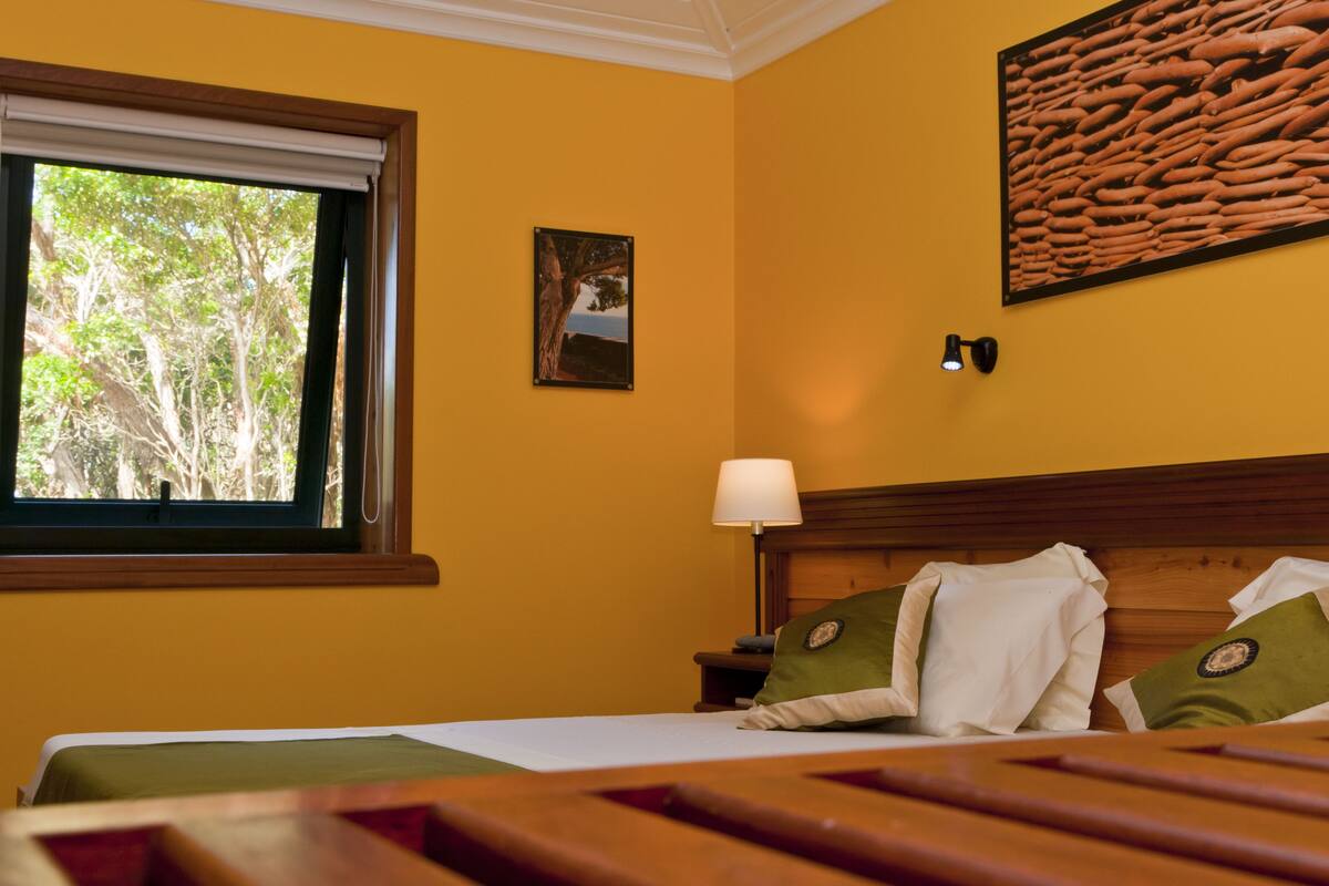 A hotel room with a bed, a window with a view of trees, a wall-mounted picture, and a wall lamp.