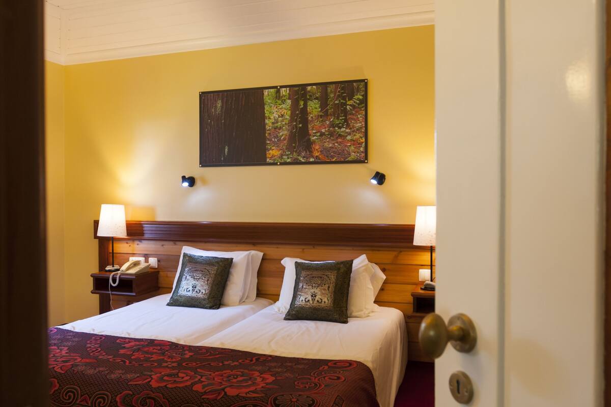 A hotel room with a double bed, two bedside lamps, a nightstand with a telephone, a painting of a forest on the wall, and a door with a lock.