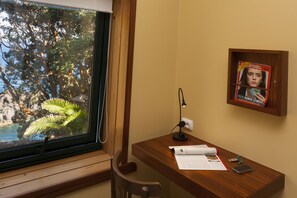 Ein Holztisch mit Lampe, Papieren und einem Mobiltelefon. Durch ein Fenster ist ein Blick auf Bäume und ein Gewässer zu sehen. Eine Zeitschrift ist an der Wand angebracht.