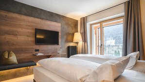 A hotel room with a large bed, a flat-screen TV mounted on a wooden wall, a bedside table with a lamp, and a window with curtains. - Christof Hotel (Monguelfo)