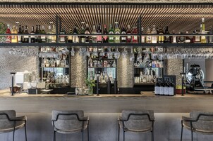 Zona de bar con varias estanterías llenas de botellas y vasos, una cafetera y asientos con sillas.