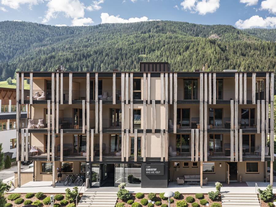A multi-story hotel with balconies, a sign reading "I AM CHRISTOF WHO ARE YOU", and a mountainous backdrop.