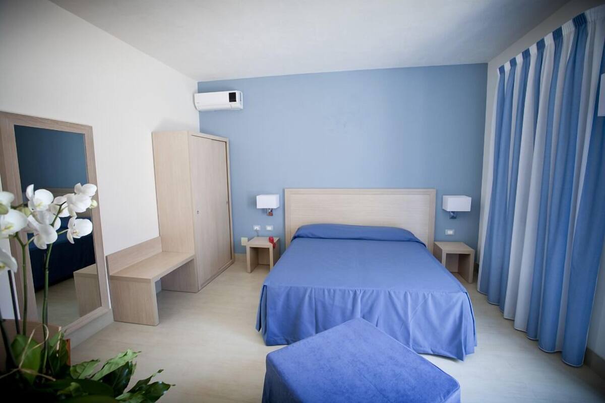 A hotel room with a single bed, a bedside table, a nightstand, a wardrobe, a mirror, and a window with curtains.