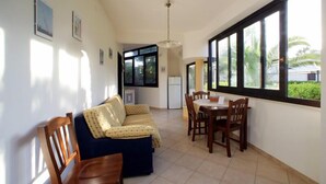 A spacious room with a sofa, dining table, and chairs. There are large windows providing natural light and a view of greenery outside. - Sunset Beach (Misiliscemi)