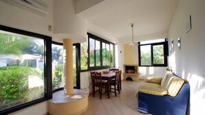 A spacious living area with a dining table, chairs, a sofa, and large windows offering a view of greenery. - Sunset Beach (Misiliscemi)