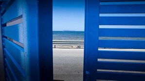 A view through a blue-framed window overlooking a beach with a concrete walkway and a distant island. - Sunset Beach (Misiliscemi)