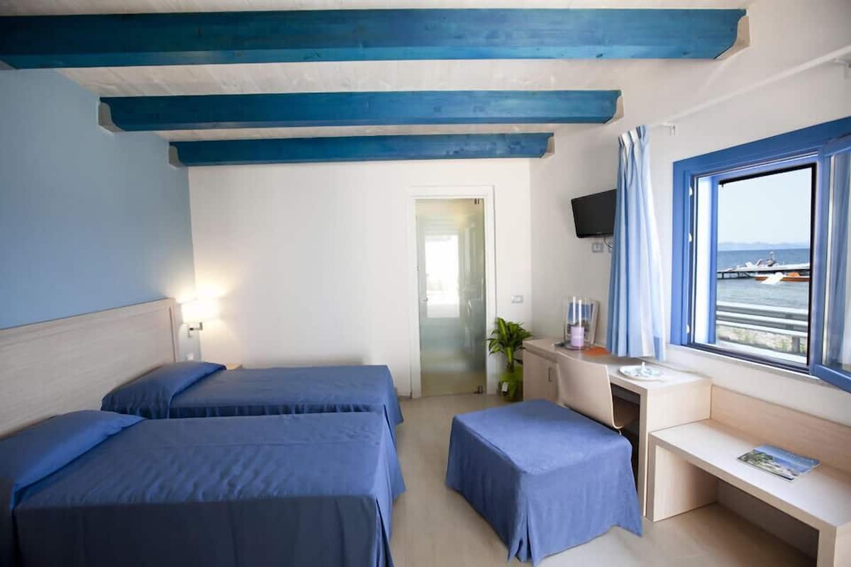 A room with two beds, a desk, a window with a view of the sea, and a TV mounted on the wall.