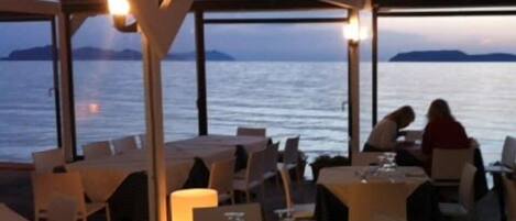 Un área de comedor al aire libre con mesas y sillas, con vista a una masa de agua y tierras distantes.