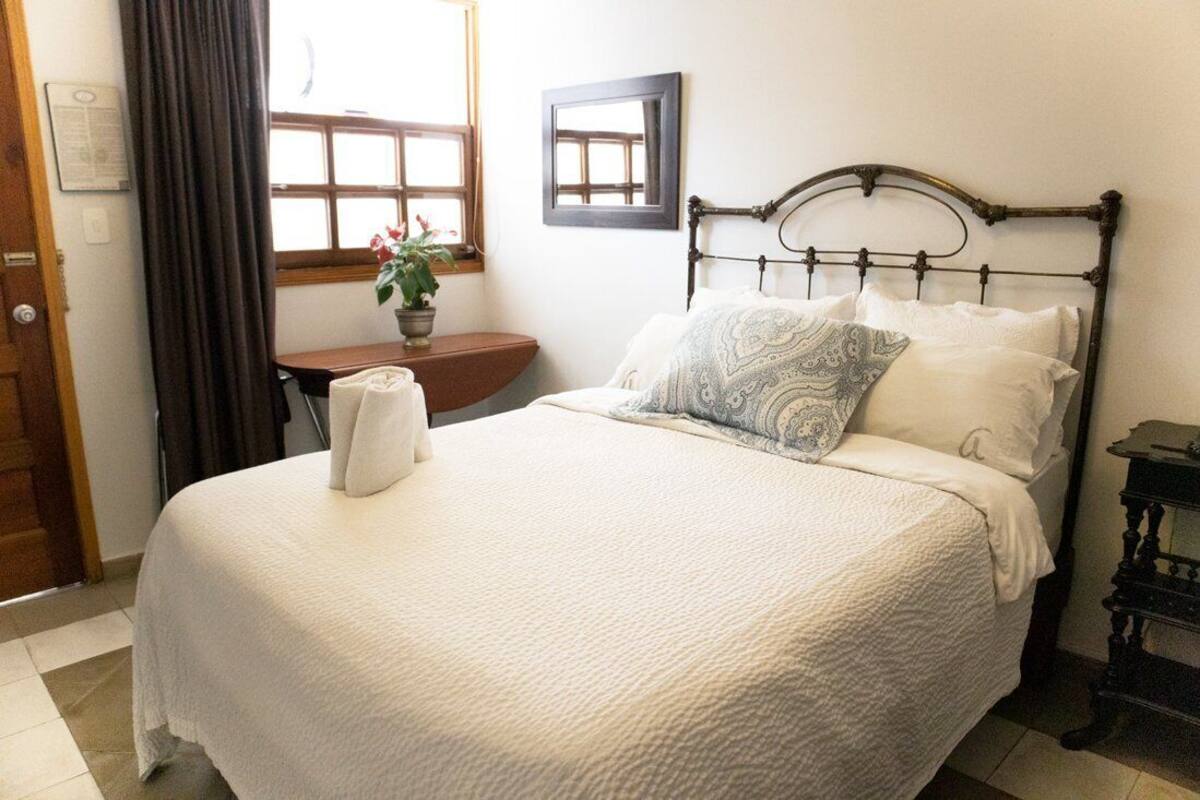A neatly made bed with a white comforter and pillows, a wooden nightstand with a lamp, and a window with curtains.