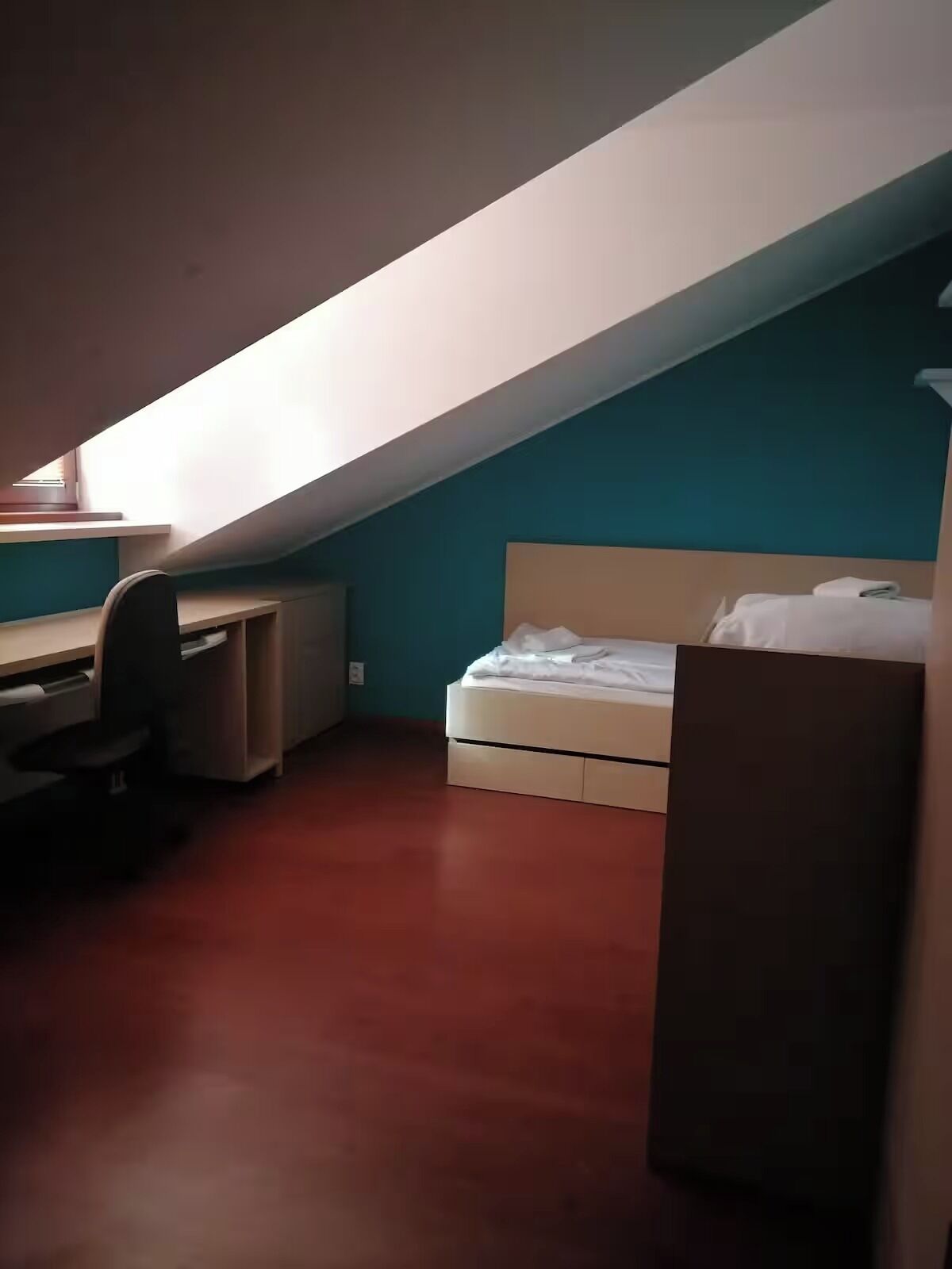 A small, dimly lit room with a sloped ceiling, a desk with a chair, and a bed.