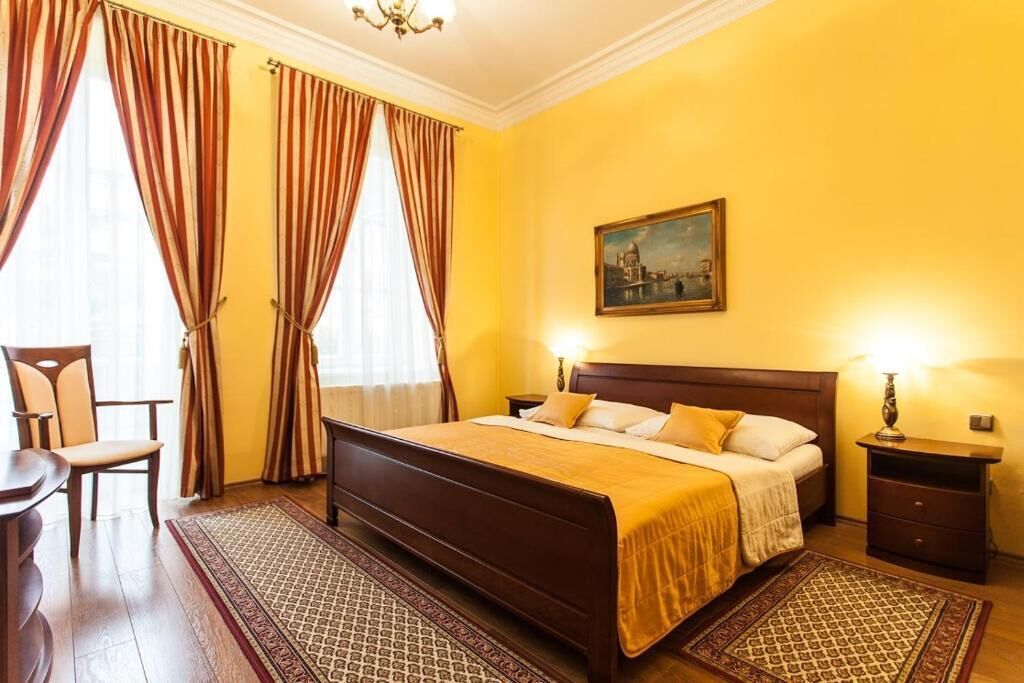 Un cuarto de hotel con una cama grande, cabecera de madera, dos mesitas de noche, una silla y un cuadro en la pared.