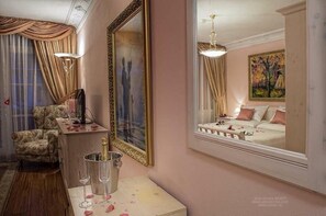 A room with a bed, a chair, a desk with a lamp, a mirror, and a painting on the wall.